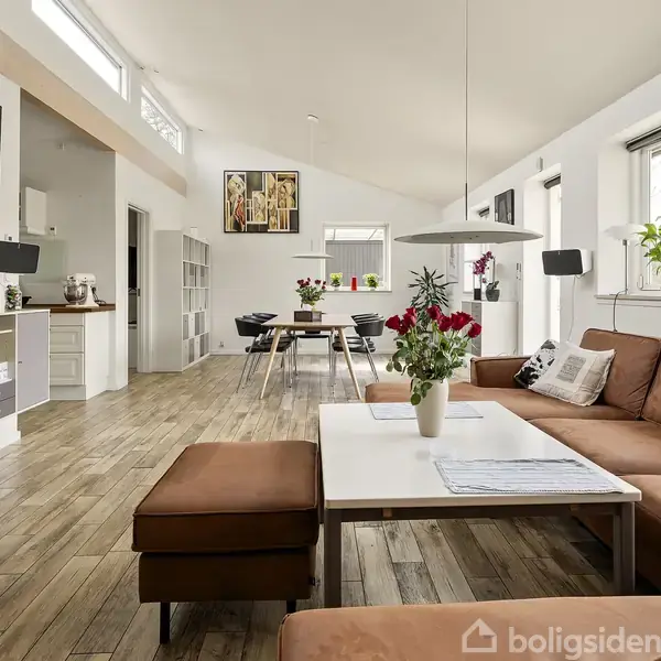 En brun lædersofa står i en moderne stue, hvor den er arrangeret omkring et hvidt sofabord med en buket blomster. Rummet er lyst med trægulve og mange vinduer.