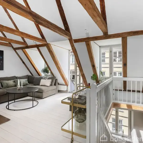 Sofagruppe i en lys stue med træbjælker i loftet, omgivet af moderne indretning. Et stort vindue giver naturligt lys, mens planter og kunst pryder rummet.