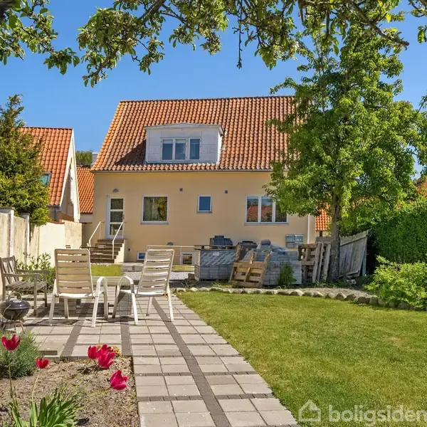 Hus med gul facade og rødt tegltag står i en grøn have. Foran huset står havemøbler på en stenbelagt sti omgivet af blomstrende tulipaner og frodig vegetation.