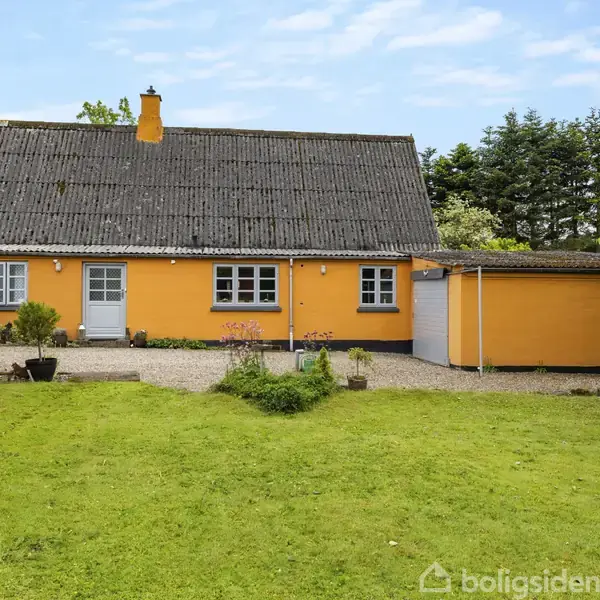 Et gult hus med sort tag står stille i en grøn have. Til venstre er der blomster og buske, mens træer omkranser baggrunden.