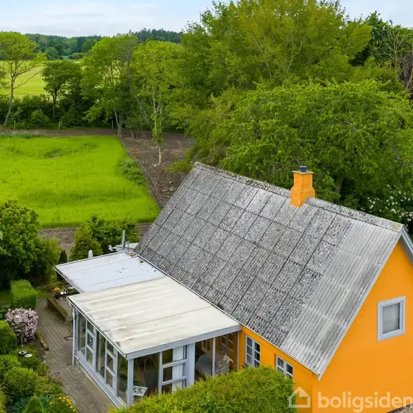 Et gult hus med skråtag står stille i en frodig have omgivet af grønne træer og marker på en solrig dag.