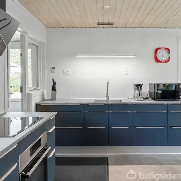 Et moderne køkken med mørkeblå skabe, hvid bordplade, og en stålvask under en væghængt lampe. Der er en rød vægur, kaffemaskine, og knivsæt. En åben dør ses til højre.