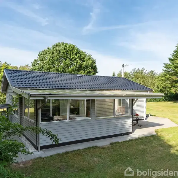 Et lille, gråt træhus med vinduer rundt om det hele står på en flisebelagt terrasse, omgivet af en grøn have med høje træer og en blå himmel i baggrunden.