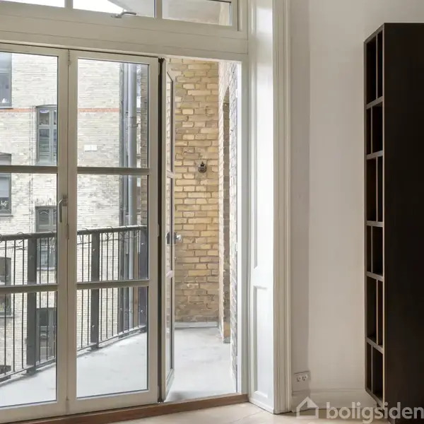 En åben altandør leder ud til en lille altan med sort rækværk. I baggrunden ses en murstensbygning.