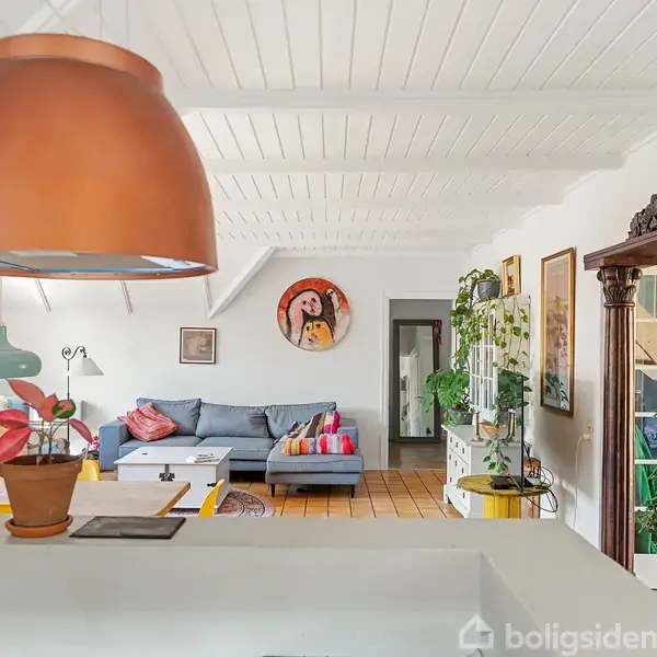 En stue med en stor, kobberfarvet lampe hængende over en køkkenø. En grå sofa med farverige puder står i rummet, omgivet af planter og kunst på væggene.