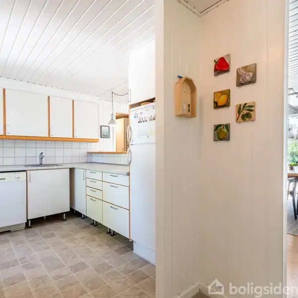 Et hvidt køkken med lyse skabe og grå flisegulv indeholder en opvaskemaskine og vask i et hjørne. I baggrunden ses et spiseområde med glasvægge. Vægge har små dekorationer.