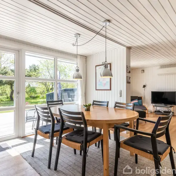 Rundt spisebord med seks stole står i et lyst rum. Store vinduer slipper sollys ind, og man kan se en grøn have udenfor. Rummet har trægulv og moderne indretning.
