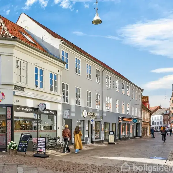 En gade med en række butikker, herunder en blomsterbutik "O.N. & KOJEF BLOMSTER" og en café med udendørs siddepladser. Folk går eller står på den brostensbelagte vej.