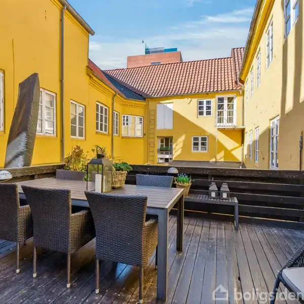 Et udendørs gårdrum med gult murværk omgiver et træbord med seks kurvestole. Plantekasser med grønne planter dekorerer omgivelserne under en klar blå himmel.