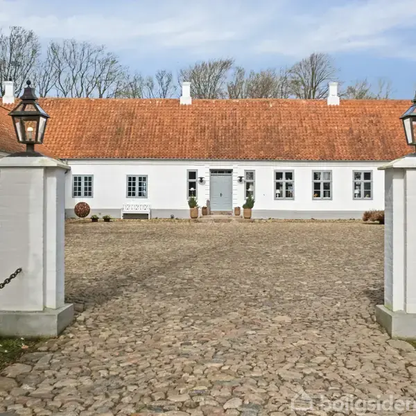 En hvid bygning med rødt tegltag står i centrum. To hvide søjler med lamper markerer en brostensbelagt indkørsel. Baggrunden domineres af træer og blå himmel.