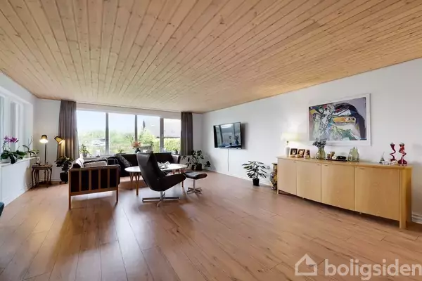 Stue møbleret med sofaer og en lænestol ved et stort vindue, fladskærm på væggen, og en trækommode med billeder og dekorationer. Lyst trægulv og loft.