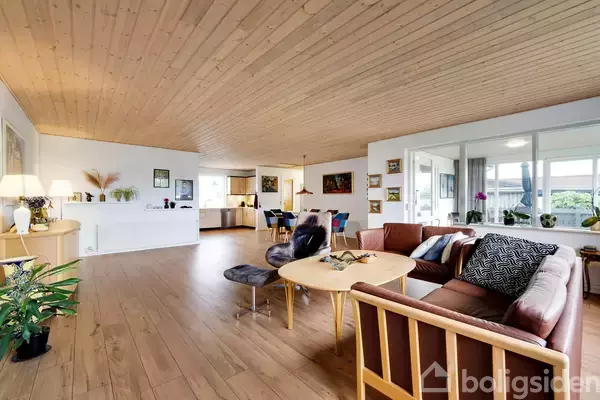 En stue med træloft har en sofagruppe centreret omkring et træsofabord. Rummet er lyst og åbent, med spisestue og køkken i baggrunden. Planter og kunst pryder væggene.