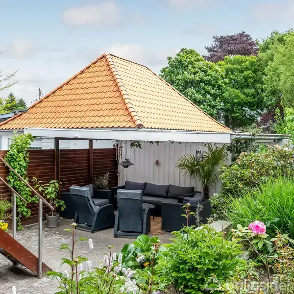 Havepavillon med rød tegltag står i en frodig have. Indenfor er der sorte loungemøbler med puder. Trætrappe ved siden af fører opad. Omkring er farverige buske og træer.