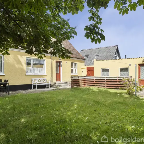 Et gult hus med en grøn græsplæne foran, omgivet af træer og buske. Der står et bord med stole på terrassen. Huset har røde døre og hvide vinduer.
