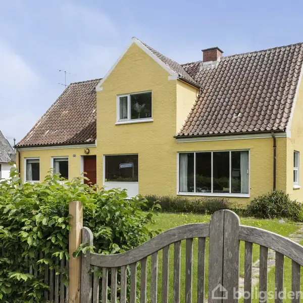 Gult murstenshus med rødt tag, store vinduer og en træport foran. Huset er omgivet af en velplejet have med buske og græsplæne.