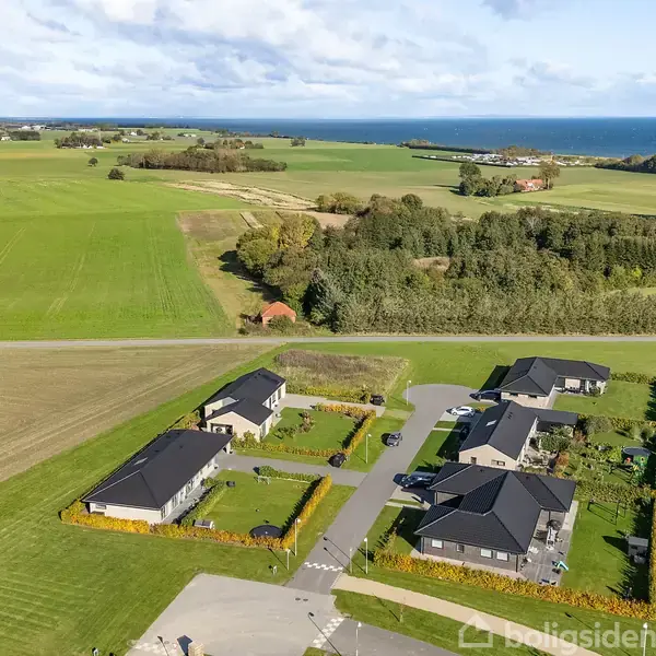 Flere huse står ud mod åbne marker og havudsigt. Husene er omgivet af velplejede haver og grusveje, med grønne marker og skov i baggrunden.