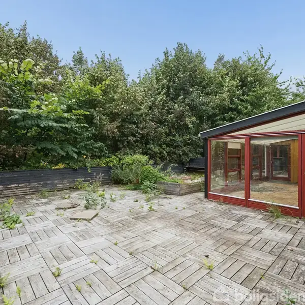 Vinterhave med røde rammer står til højre, omgivet af et stort terrasseområde med kvadratiske fliser og spredte planter. I baggrunden findes en tæt bevoksning af buske og træer.