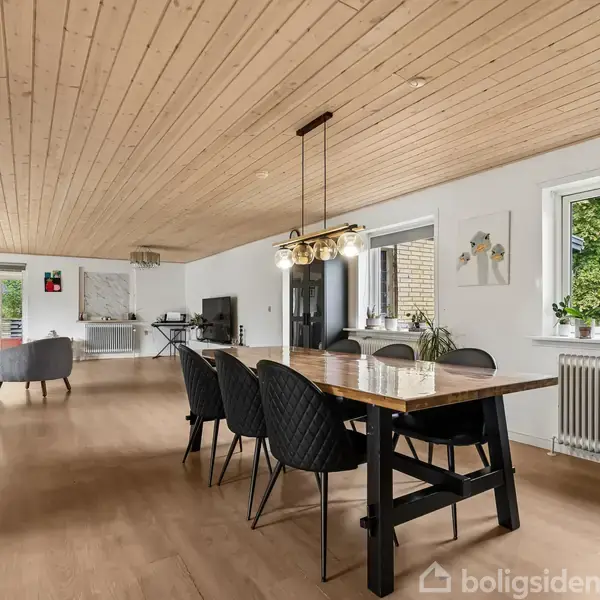Spisebord står i centrum med sorte stole omkring, under en lampe. Rummet har trægulv og træloft, store vinduer med udsigt til grønne træer, og moderne indretning med sofa i baggrunden.