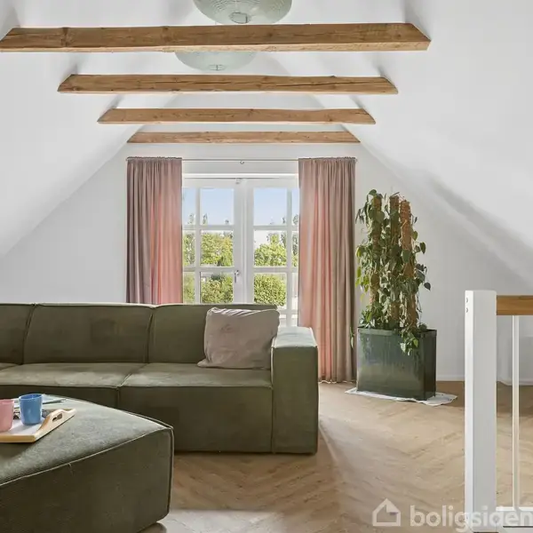 En grøn sofa står i en lys stue under træbjælker, nær en vindue med lyserøde gardiner. På sofabordet er der en bakke med kopper. En plante står ved siden.