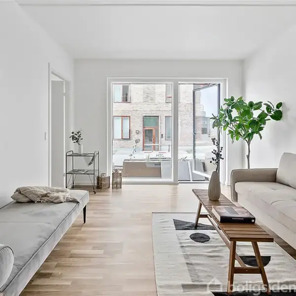 En lys stue indeholder en beige sofa og et sofabord, der står på et trægulv. En stor plante står i hjørnet nær en stor glasdør. To små billeder hænger på væggene.