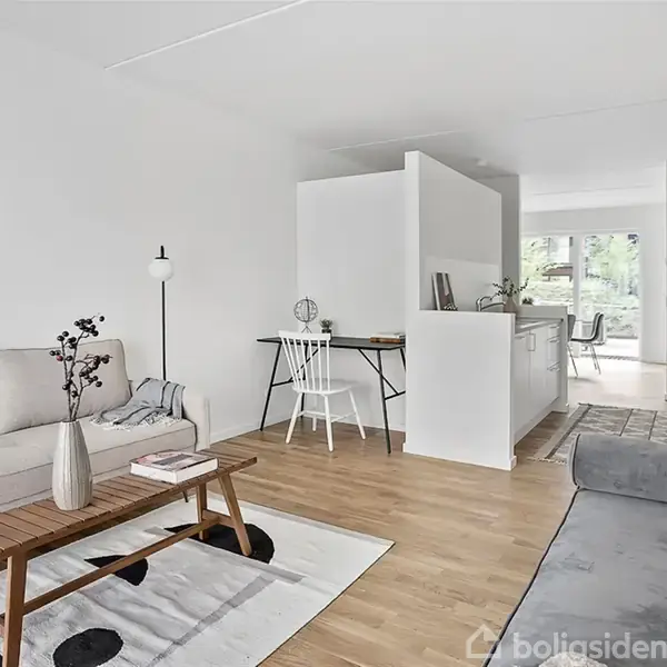 En lys stue indeholder en sofa, et lille træbord med vase og bøger, og en kontorplads i baggrunden. Rummets indretning er moderne og minimalistisk.
