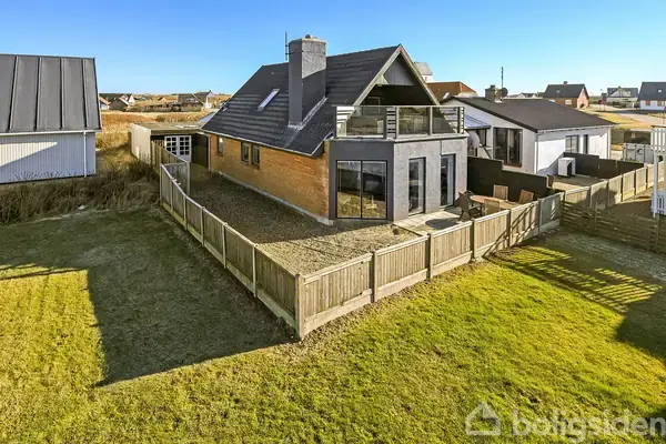 Hus med terrasse og balkon, står i en indhegnet have med græsplæne. Det er omgivet af andre huse i et boligområde under en klar blå himmel.