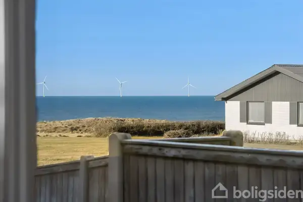 Hus set fra en terrasse med udsigt til havet og flere vindmøller i horisonten. Foran verandaen er der hegn og en græsplæne, mens himlen er klar og blå.