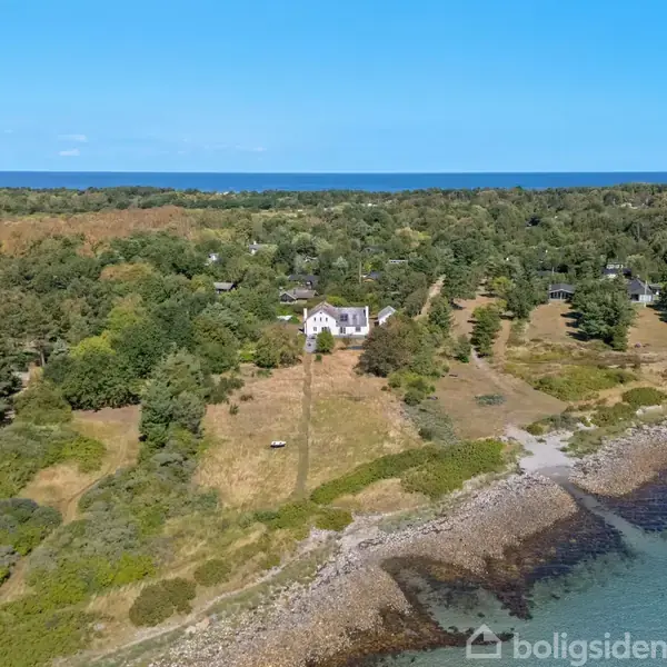 Huset ligger midt i en skovklædt kystlinje, med udsigt over en stenstrand og havet i baggrunden. Omkringliggende grønne og tætte træer skaber en rolig atmosfære.