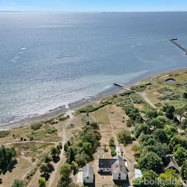 Et hus står stille ved kysten omgivet af træer og grønne områder. Havet strækker sig i baggrunden med moler, der rækker ud i vandet under en klar himmel.