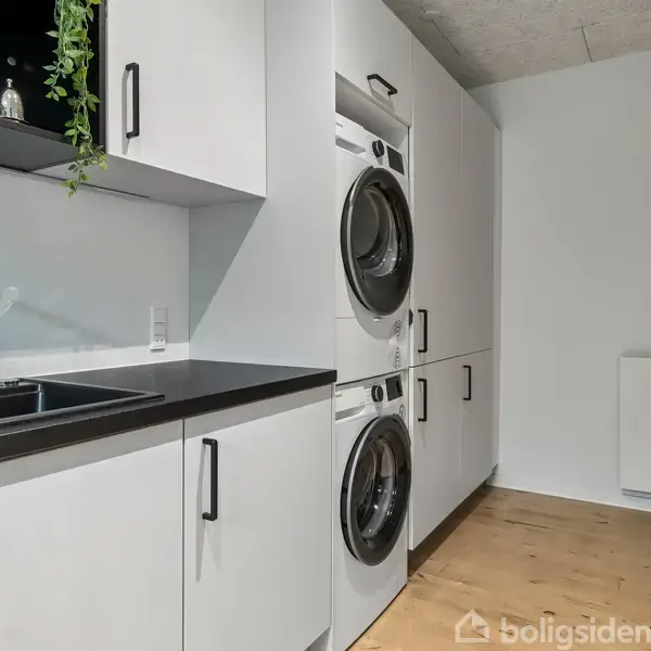 Vaskemaskine og tørretumbler er stablet i et moderne bryggers med hvide skabe. Der er en sort håndvask på en sort bordplade. Lyse trægulve fuldender rummet.