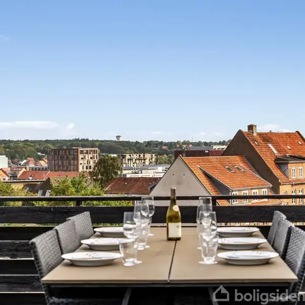 Et udendørs bord dækket med tallerkener, glas og en flaske vin står på en balkon med udsigt over hustage i en by.