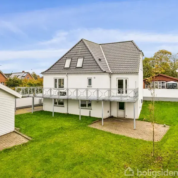 Hus med hvid facade og gråt tag står i en grøn have med en træterrasse. Nabohuse kan ses i baggrunden.