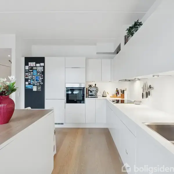 Et moderne køkken i hvidt design med trælaminatgulv. En rød vase med blomster står på en køkkenø. Skabe indeholder køkkenredskaber; billeder og tekst "NOT WITHOUT US" ses på væggen.