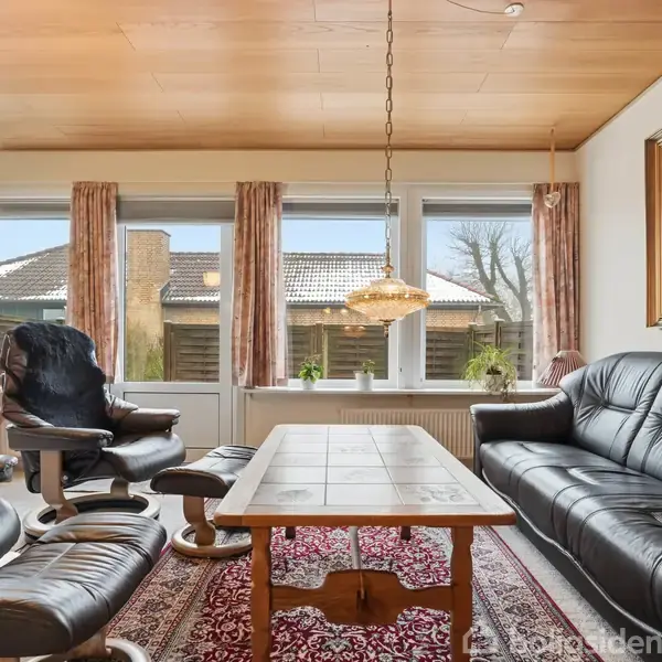 En stue indeholder sorte lædermøbler placeret omkring et rektangulært sofabord. Store vinduer med gardiner lader lys ind, og en hængelampe hænger over bordet. Væggen har et stort maleri.