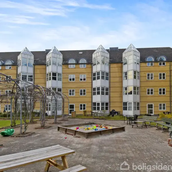 Boligkompleks med gule mursten og glasbalkoner. I forgrunden ses en legeplads med sandkasse og legetøj. Bænke og græsarealer omgiver legeområdet.