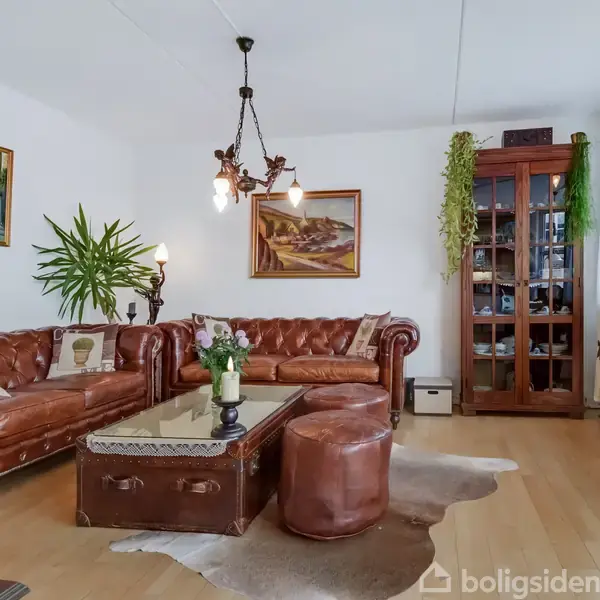 Brune lædersofaer står i en elegant stue med trægulve og kunst på væggene. Et sofabord med stearinlys står i midten, mens en glasvitrine og planter pynter rummet.