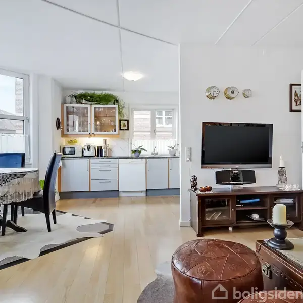 Stue med fjernsyn på tv-bord, lænestol i forgrunden, spisebord ved vindue, åbent køkken i baggrunden med hvide skabe og planter ovenpå. Lyst rum med lyse trægulve.