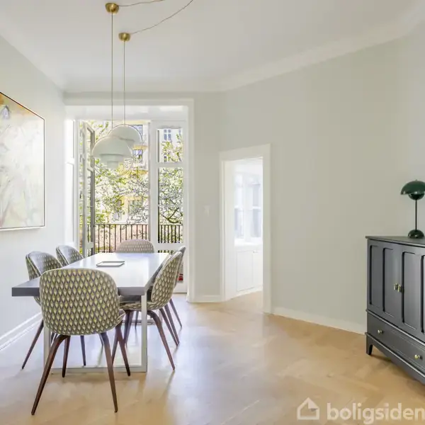 Spisebord med seks stole står i et lyst rum med stor vinduesparti, der åbner ud mod natur. Et maleri hænger på væggen, og en kommode med lampe står i hjørnet.