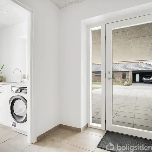 En vaskemaskine og tørretumbler står ved siden af et hvidt skab med en vask. Gennem glasdøre ses en garage med parkerede biler udenfor på terrassen.