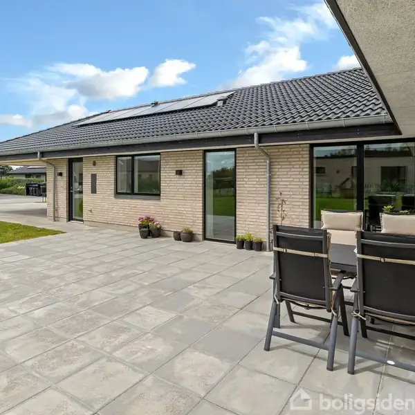 Moderne hus med et fladt tag og store vinduer. Terrasse med flisebelægning og havemøbler. Omkringliggende grøn plæne og ekstra bygninger i baggrunden. Solrige og klare himmel.