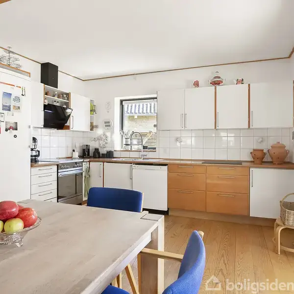 Et køkken med træ- og hvide skabe, hvor en frugtskål står på et bord med blå polstrede stole. Omkring er der en ovn, køleskab og et vindue.