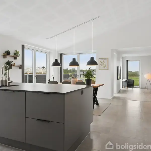 Køkkenø med vask og kogeplade står i et lyst, moderne køkken-alrum med spisebord og lamper, store vinduer og udsigt til en grøn have i baggrunden.