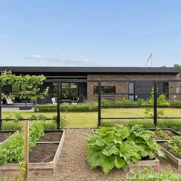 En moderne murstensvilla står i en velplejet have med højbede, hvor grøntsager vokser. Bag ved huset er en terrasse med havemøbler. Himmel og grønt landskab omgiver scenen.
