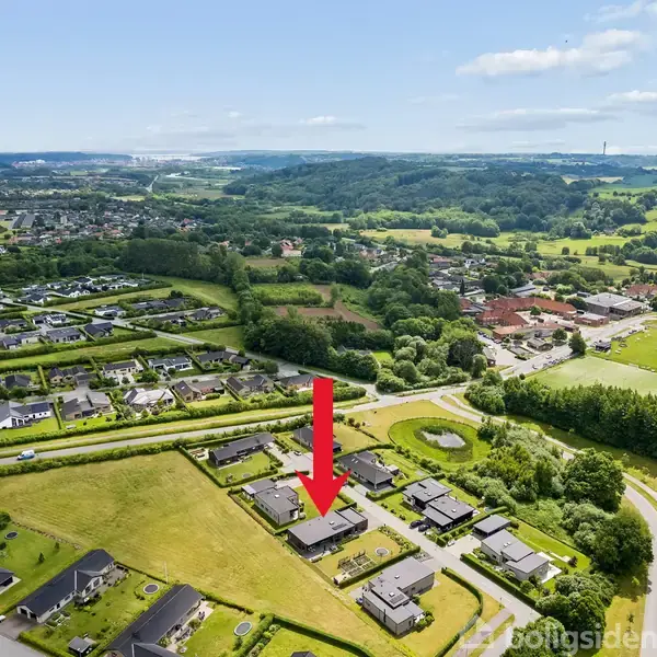 En moderne murstensvilla med fladt tag står i en velplejet, grøn have. Der er en rund trampolin i græsplænen og vej omgivet af frodige buske og træer.