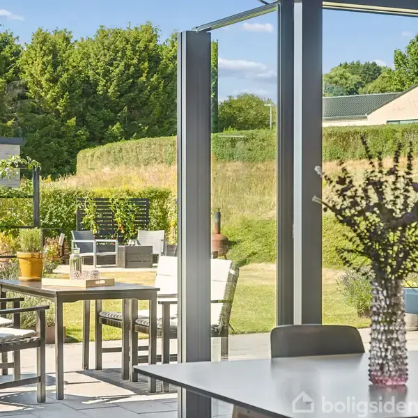 Indendørs bord med blomster i vase står ved store glasdøre, der åbner ud til en solrig terrasse med træmøbler og planter, omkranset af grønne hække og træer.