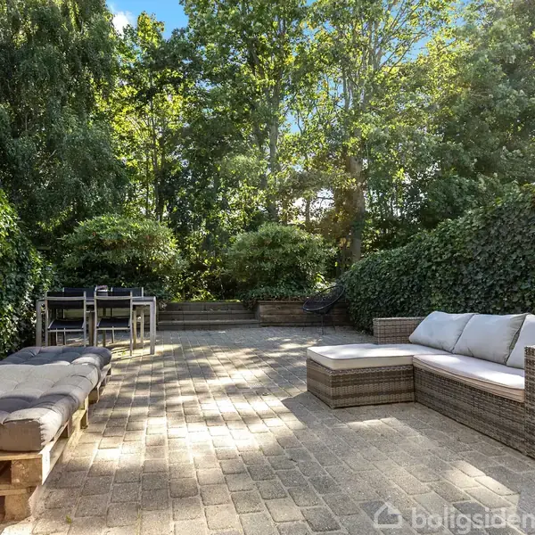 Terrassemøbler med sofa og pallebænk står på en flisebelagt terrasse omgivet af høje grønne hække og træer; et spisebord og stole ses i baggrunden.