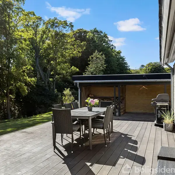 Et træterrassebord med seks stole står stille i solskin ved siden af et hus. I baggrunden ses en have med grønne træer under en klar blå himmel.