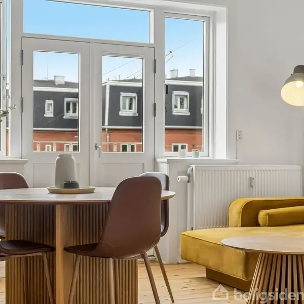 Et rundt spisebord med brune stole står i et lyst rum med store vinduer. En gul sofa og en tændt lampe ses i baggrunden.