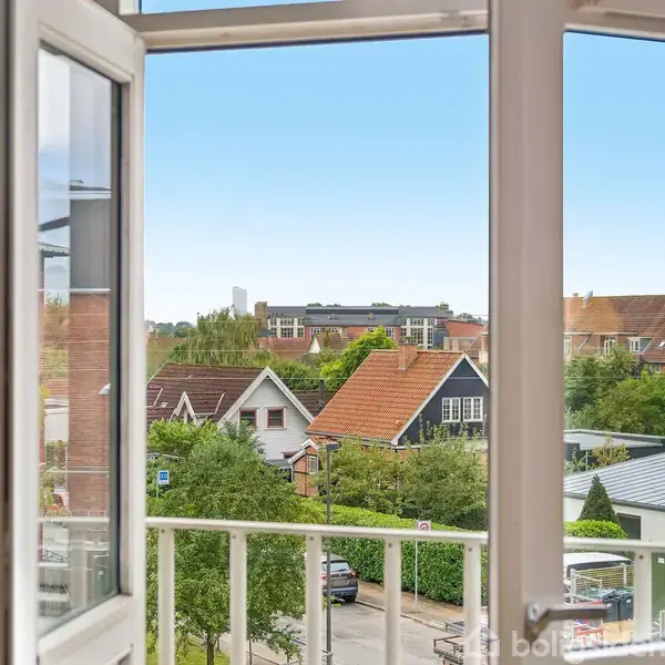 Åbent vindue afslører udsigt til et villakvarter; huse med røde tage omkranset af grønne træer. Blå vase med blomster står i vindueskarmen.