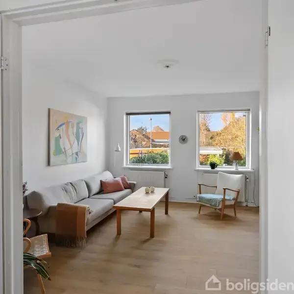 Stue med sofa og træbord, placeret ved store vinduer, der giver udsigt til en solrig have. Vægmaleri hænger over sofaen, og en gyngestol står ved vinduet.